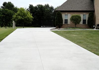 A clean, wide concrete driveway leads to a house with neatly trimmed bushes and a green lawn, resembling a stunning display in a paving gallery. Trees are in the background.