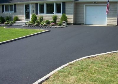 residential asphalt companies A freshly paved asphalt driveway leading to a single-story house with a green lawn and an American flag, akin to a pristine exhibit in a paving gallery.