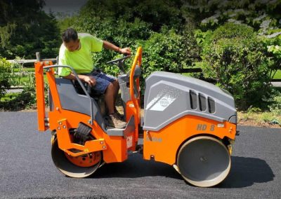 Construction worker operating an orange road roller on a freshly paved, black asphalt surface with greenery in the background, showcasing the scene like a live paving gallery.
