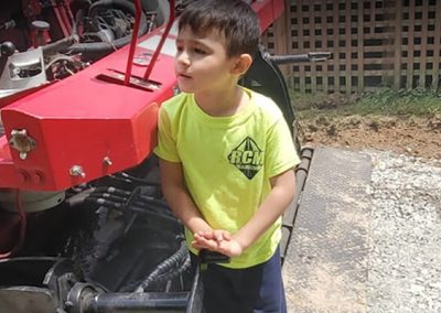 A young boy in a yellow shirt stands by a red tractor, looking to the side, with a lattice structure and paving in the background.