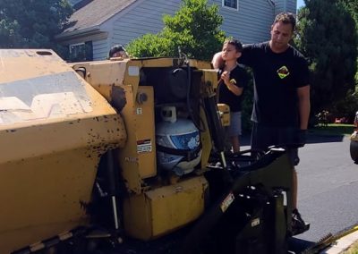 Workers stand near a large yellow paving machine, possibly for roadwork, on a sunny day in a residential area.