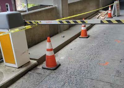 A parking garage entrance blocked with traffic cones, caution tape, and a barrier arm. Construction materials are scattered in the background, turning the site into a makeshift paving gallery.