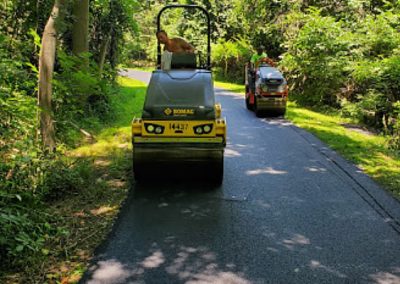 Two road rollers paving an asphalt path through a forested area with greenery on both sides, creating a scenic paving gallery.