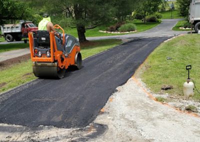 A worker on a roller compactor smoothing a newly paved asphalt driveway in a residential area, contributing to the neighborhood's paving gallery of pristine roads.