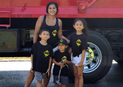 A woman and three children in matching shirts and hats pose cheerfully in front of a large truck, creating a charming family moment reminiscent of a gallery exhibition.