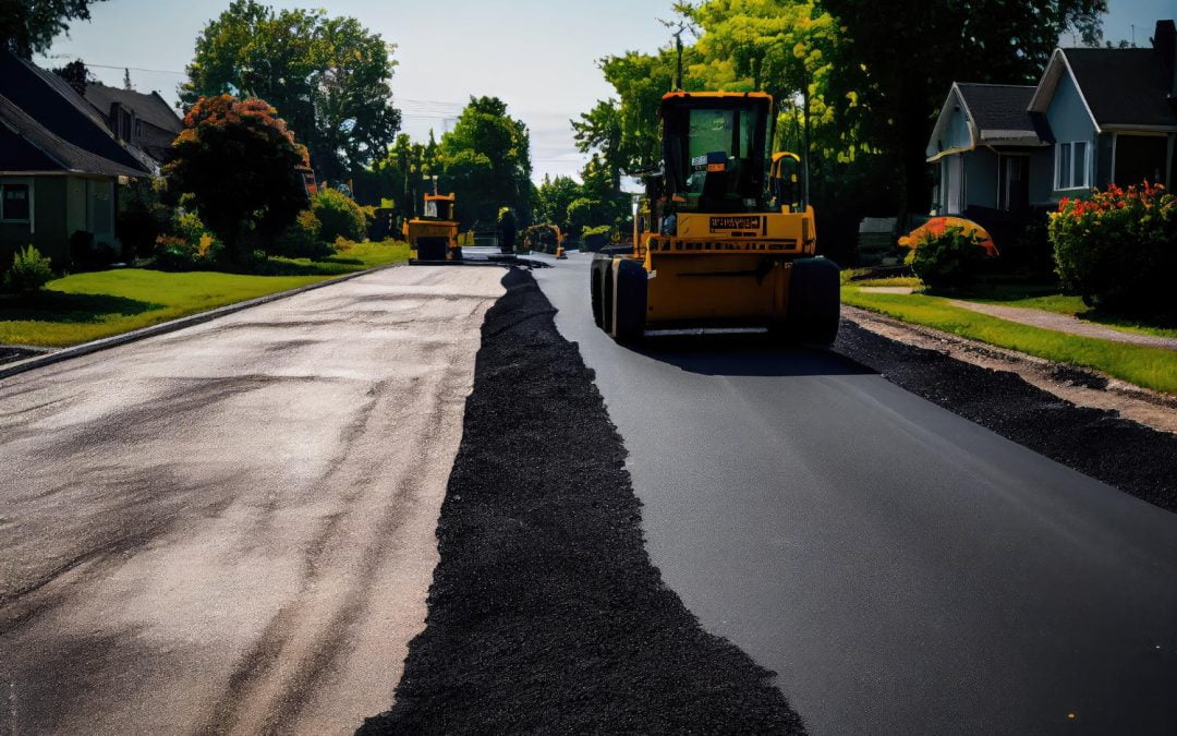 Road construction with heavy machinery paving a residential street, creating smooth asphalt driveways. Trees and houses are visible on both sides of the road.