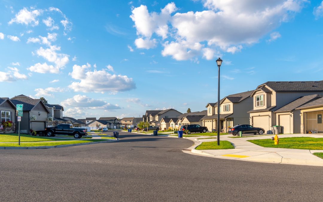 Suburban neighborhood with modern houses, cars parked in driveways, and a clear blue sky with scattered clouds. Ongoing road projects are subtly enhancing the charm and accessibility of this idyllic setting.