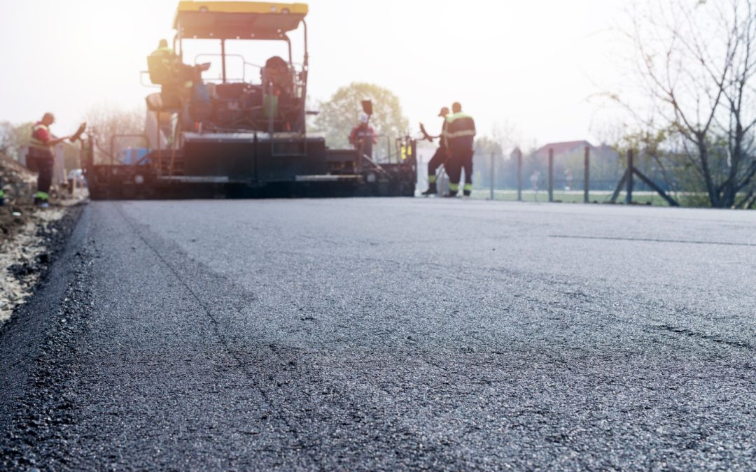 Workers paving a road with machinery on a sunny day, with fresh asphalt in the foreground.